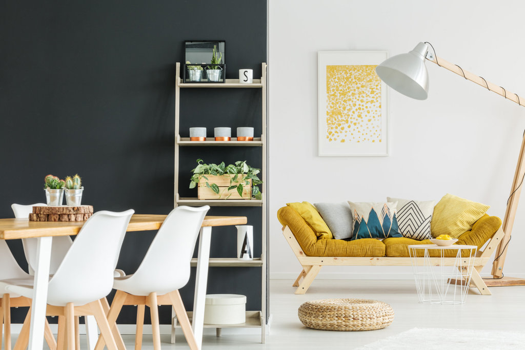 2 stanze sono delineate dal colore delle pareti. a destra, si può vedere il soggiorno con uno stile di legno e giallo su una parete bianca. a sinistra, si può indovinare la cucina su una parete nera con un tavolo di legno in stile scandinavo e sedie bianche.