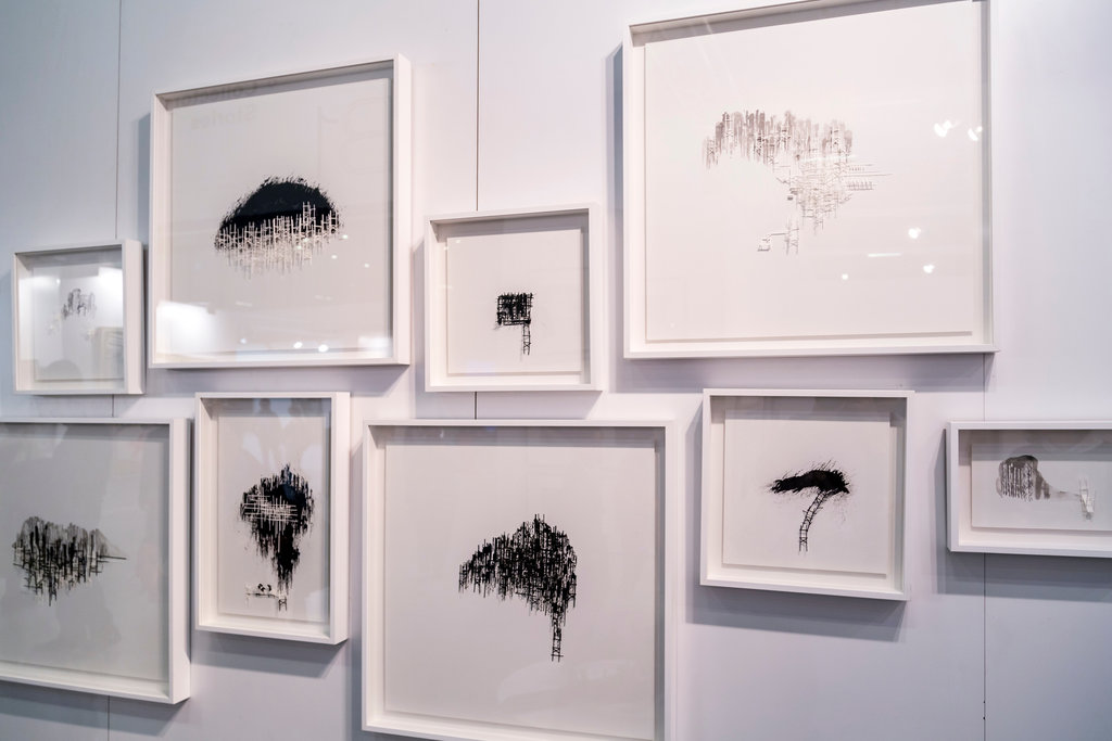 Sie sehen eine Reihe von Werken in Schwarz auf weißem Hintergrund, gerahmt in Glasrahmen mit weißen Rändern und ausgestellt an einer weißen Wand