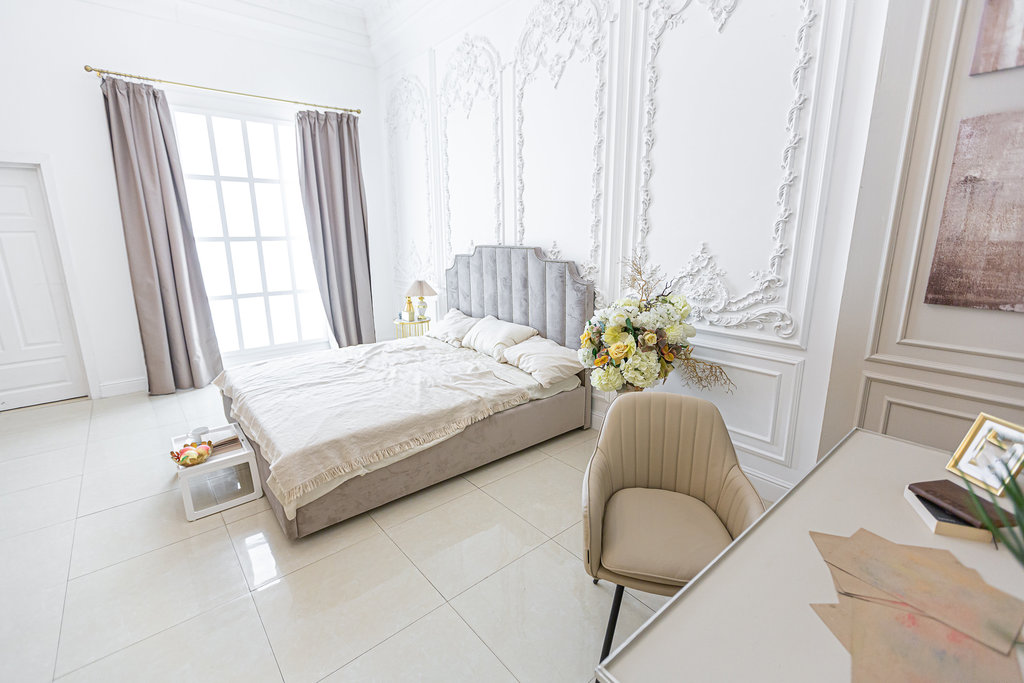 Zeer ruime oude chique kamer ingericht met lijstwerk op de witte muren, een nep groot boeket bloemen bij het bed, wit, taupe en beige kleuren
