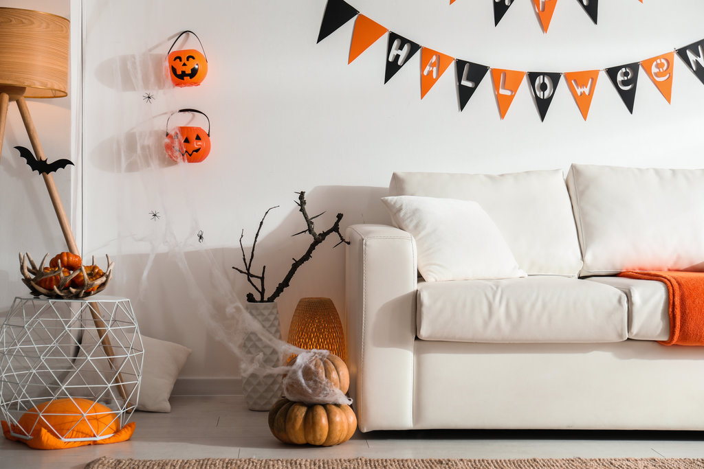 ein weißes wohnzimmer ist mit schwarzer und orangefarbener halloween-dekoration geschmückt. eine fröhliche halloween-girlande hängt über dem weißen sofa, während kürbisse an der wand hängen und auf dem boden liegen. an den kürbissen hängt ein spinnennetz