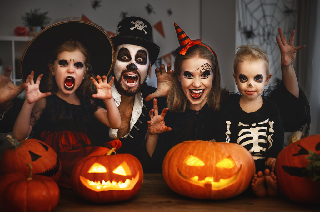 eine familie, bestehend aus papa, mama und ihren 2 kindern (mädchen und junge), macht mit offenem mund eine gruselige pause, verkleidet für halloween. vor ihnen liegen beleuchtete und furchterregende kürbisse auf dem tisch.