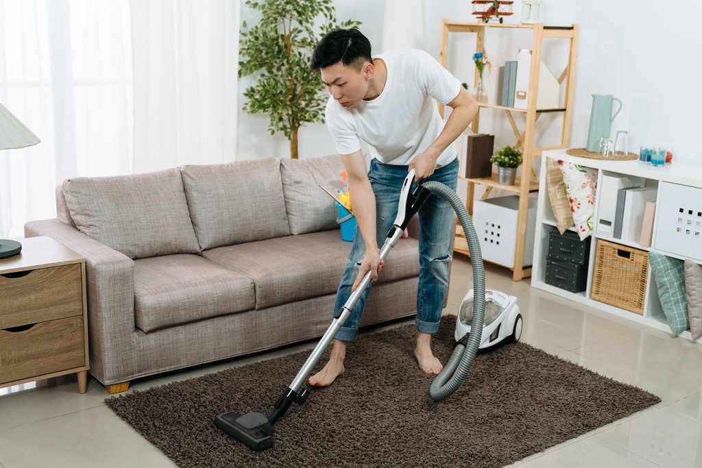 een man, van Aziatische afkomst, is te zien terwijl hij zijn woonkamer stofzuigt