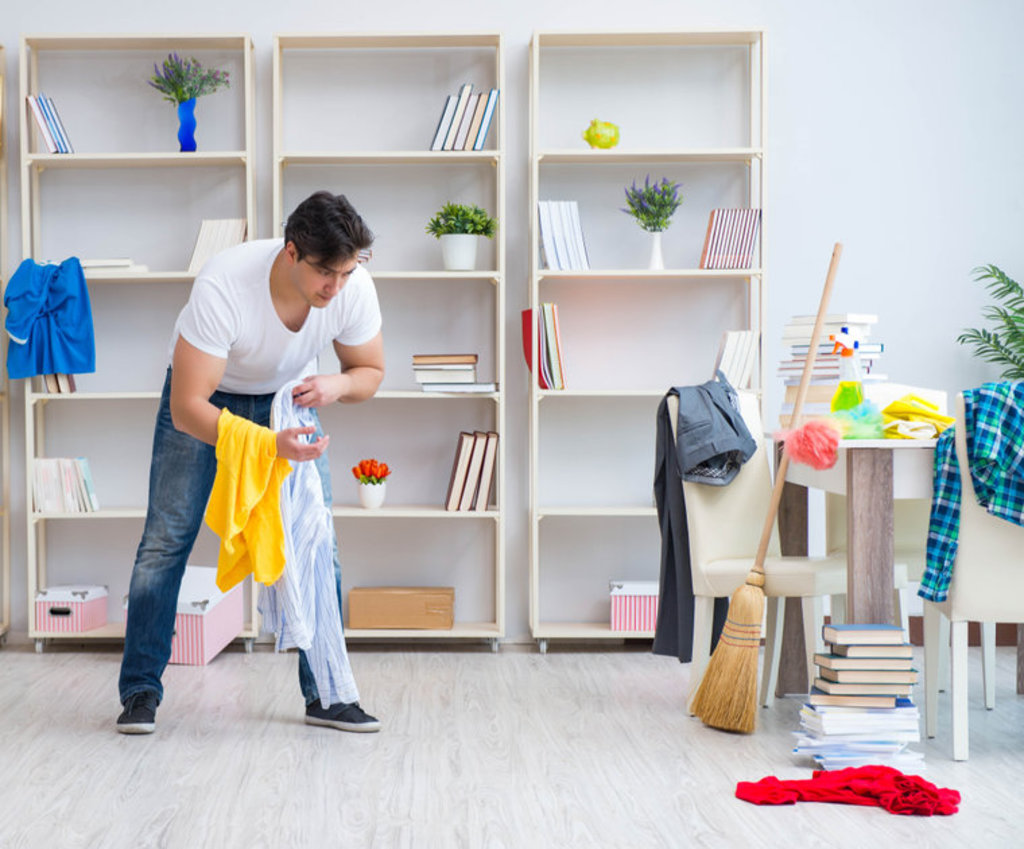 een man in een wit t-shirt en spijkerbroek, raapt kleren op die in zijn woonkamer liggen. een bezem staat tegen de tafel geleund omdat het tijd is om op te ruimen en schoon te maken.